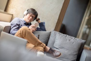 Genç Asyalı adam kanepede smartphone kullanarak müzik dinlemek