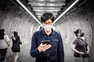 Asyalı bir adam metro tünelinde kalabalık insanların yürüdüğü akıllı telefon kullanarak cerrahi maske takıyor. Wuhan Coronavirus (Covid-19) kamu alanındaki salgın önleme. Sağlık ve tıbbi konsept