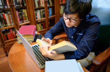 Asyalı adam üniversitesi kitapları okuyor ve kolej kütüphanesinde kitaplığın yanında dizüstü bilgisayar kullanıyor. Eğitim araştırmaları için ders kitabı kaynakları. Burs fırsatı kavramı