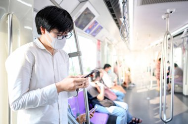 Asyalı bir adam hava treninde ya da şehir treninde akıllı telefon kullanarak cerrahi maske takıyor. Wuhan Coronavirus (COVID-19) toplu taşımacılıkta salgın önleme. Pandemik koruma için sağlık farkındalığı
