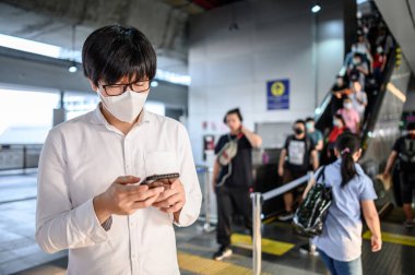 Skytrain istasyonunda akıllı telefonu kullanarak cerrahi maske takan Asyalı bir adam. Wuhan Coronavirus (COVID-19) toplu taşımacılıkta salgın önleme. Pandemik koruma için sağlık farkındalığı