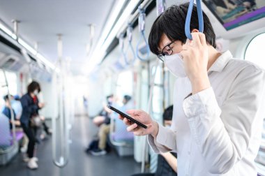 Asyalı bir adam hava treninde ya da şehir treninde akıllı telefon kullanarak cerrahi maske takıyor. Wuhan Coronavirus (COVID-19) toplu taşımacılıkta salgın önleme. Pandemik koruma için sağlık farkındalığı