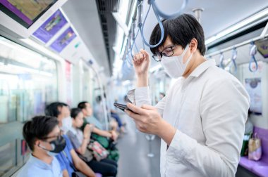 Asyalı bir adam hava treninde ya da şehir treninde akıllı telefon kullanarak cerrahi maske takıyor. Wuhan Coronavirus (COVID-19) toplu taşımacılıkta salgın önleme. Pandemik koruma için sağlık farkındalığı