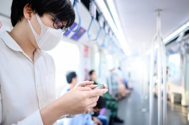 Asyalı bir adam hava treninde ya da şehir treninde akıllı telefon kullanarak cerrahi maske takıyor. Wuhan Coronavirus (COVID-19) toplu taşımacılıkta salgın önleme. Pandemik koruma için sağlık farkındalığı