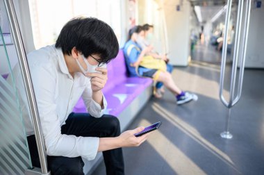 Asyalı bir adam hava treninde ya da şehir treninde akıllı telefon kullanarak cerrahi maske takıyor. Wuhan Coronavirus (COVID-19) toplu taşımacılıkta salgın önleme. Pandemik koruma için sağlık farkındalığı
