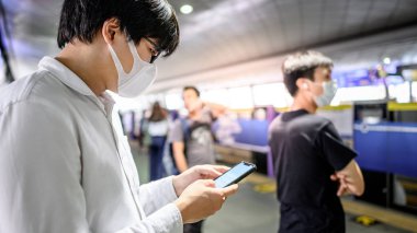 Metro istasyonunda akıllı telefon kullanarak cerrahi maske takan Asyalı bir adam. Wuhan Coronavirus (COVID-19) toplu taşımacılıkta salgın önleme. Pandemik koruma için sağlık farkındalığı