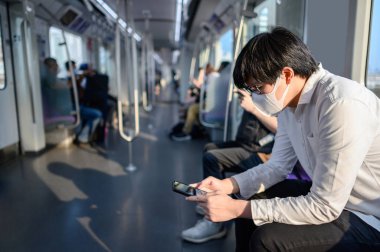 Asyalı bir adam hava treninde ya da şehir treninde akıllı telefon kullanarak cerrahi maske takıyor. Wuhan Coronavirus (COVID-19) toplu taşımacılıkta salgın önleme. Pandemik koruma için sağlık farkındalığı