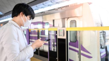 Skytrain istasyonunda akıllı telefonu kullanarak cerrahi maske takan Asyalı bir adam. Wuhan Coronavirus (COVID-19) toplu taşımacılıkta salgın önleme. Pandemik koruma için sağlık farkındalığı