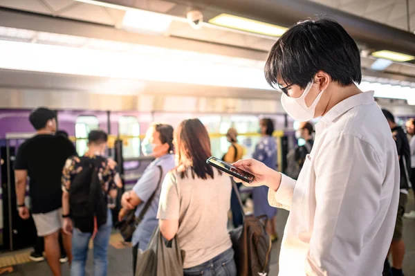 Skytrain istasyonunda akıllı telefonu kullanarak cerrahi maske takan Asyalı bir adam. Wuhan Coronavirus (COVID-19) toplu taşımacılıkta salgın önleme. Pandemik koruma için sağlık farkındalığı