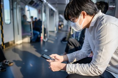 Asyalı bir adam hava treninde ya da şehir treninde akıllı telefon kullanarak cerrahi maske takıyor. Wuhan Coronavirus (COVID-19) toplu taşımacılıkta salgın önleme. Pandemik koruma için sağlık farkındalığı