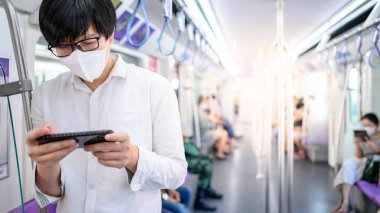 Asyalı bir adam hava treninde ya da şehir treninde akıllı telefon kullanarak cerrahi maske takıyor. Wuhan Coronavirus (COVID-19) toplu taşımacılıkta salgın önleme. Pandemik koruma için sağlık farkındalığı