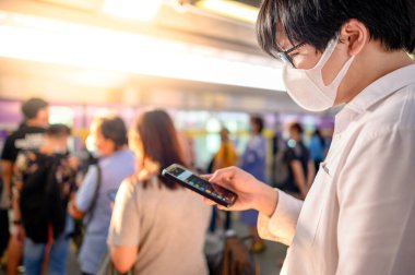 Skytrain istasyonunda akıllı telefonu kullanarak cerrahi maske takan Asyalı bir adam. Wuhan Coronavirus (COVID-19) toplu taşımacılıkta salgın önleme. Pandemik koruma için sağlık farkındalığı
