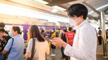Skytrain istasyonunda akıllı telefonu kullanarak cerrahi maske takan Asyalı bir adam. Wuhan Coronavirus (COVID-19) toplu taşımacılıkta salgın önleme. Pandemik koruma için sağlık farkındalığı