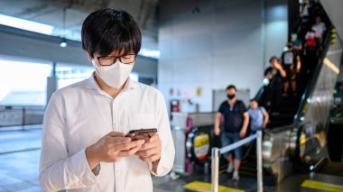 Skytrain istasyonunda akıllı telefonu kullanarak cerrahi maske takan Asyalı bir adam. Wuhan Coronavirus (COVID-19) toplu taşımacılıkta salgın önleme. Pandemik koruma için sağlık farkındalığı