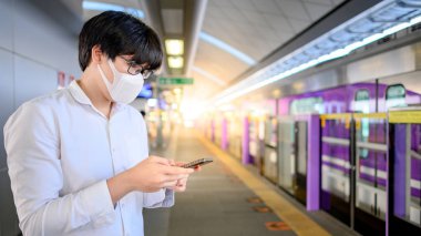 Skytrain istasyonunda akıllı telefonu kullanarak cerrahi maske takan Asyalı bir adam. Wuhan Coronavirus (COVID-19) toplu taşımacılıkta salgın önleme. Pandemik koruma için sağlık farkındalığı