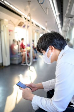 Asyalı bir adam hava treninde ya da şehir treninde akıllı telefon kullanarak cerrahi maske takıyor. Wuhan Coronavirus (COVID-19) toplu taşımacılıkta salgın önleme. Pandemik koruma için sağlık farkındalığı