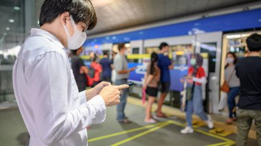 Skytrain istasyonunda akıllı telefonu kullanarak cerrahi maske takan Asyalı bir adam. Wuhan Coronavirus (COVID-19) toplu taşımacılıkta salgın önleme. Pandemik koruma için sağlık farkındalığı