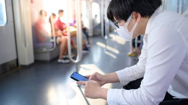 Asyalı bir adam hava treninde ya da şehir treninde akıllı telefon kullanarak cerrahi maske takıyor. Wuhan Coronavirus (COVID-19) toplu taşımacılıkta salgın önleme. Pandemik koruma için sağlık farkındalığı