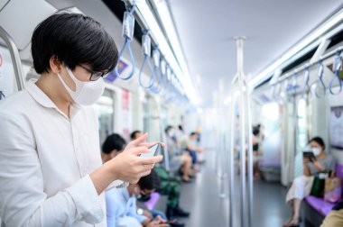 Asyalı bir adam hava treninde ya da şehir treninde akıllı telefon kullanarak cerrahi maske takıyor. Wuhan Coronavirus (COVID-19) toplu taşımacılıkta salgın önleme. Pandemik koruma için sağlık farkındalığı