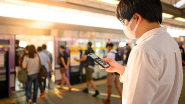Skytrain istasyonunda akıllı telefonu kullanarak cerrahi maske takan Asyalı bir adam. Wuhan Coronavirus (COVID-19) toplu taşımacılıkta salgın önleme. Pandemik koruma için sağlık farkındalığı