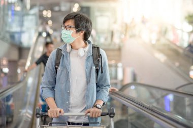 Havaalanı terminalinde havaalanı tramvayı ve bavuluyla yürüyen Asyalı adam yüz maskesi takıyor. Yurtdışına seyahat ederken Coronavirus (Covid-19) salgını önleme. Pandemik koruma için sağlık farkındalığı