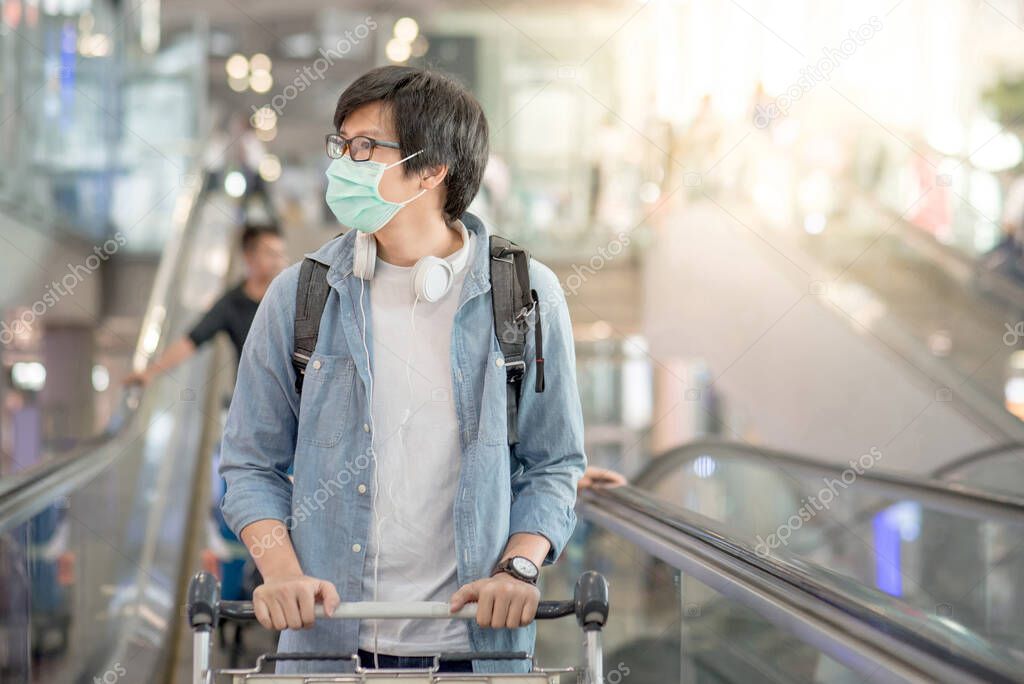 Asian man wear face mask walking with airport trolley and suitcase luggage in airport terminal. Coronavirus (COVID-19) outbreak prevention when travel abroad. Health awareness for pandemic protection