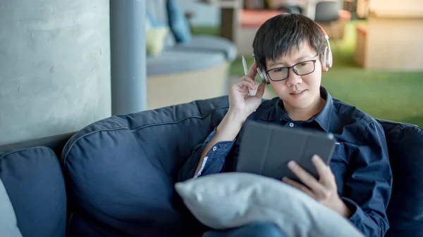 Asian businessman with headphones listening to music working with digital tablet on comfortable sofa at home office. Man with glasses doing online conference on mobile app. Work from home concept