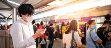 Skytrain istasyonunda akıllı telefonu kullanarak cerrahi maske takan Asyalı bir adam. Wuhan Coronavirus (COVID-19) toplu taşımacılıkta salgın önleme. Pandemik koruma için sağlık farkındalığı