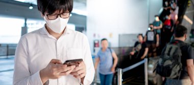 Skytrain istasyonunda akıllı telefonu kullanarak cerrahi maske takan Asyalı bir adam. Wuhan Coronavirus (COVID-19) toplu taşımacılıkta salgın önleme. Pandemik koruma için sağlık farkındalığı