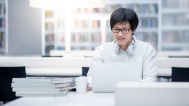 Genç Asyalı erkek üniversite öğrencisi üniversite kütüphanesinde eğitim araştırmaları için dizüstü bilgisayar kullanıyor. Erkek araştırmacı internet platformunda bilgi arıyor. Çevrimiçi öğrenim veya E-öğrenme kavramı