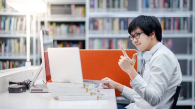 Genç Asyalı erkek üniversite öğrencisi üniversite kütüphanesinde bilgisayar ve dizüstü bilgisayar kullanıyor. Erkek araştırmacı internet platformunda bilgi topluyor. Çevrimiçi öğrenim veya E-öğrenme kavramı