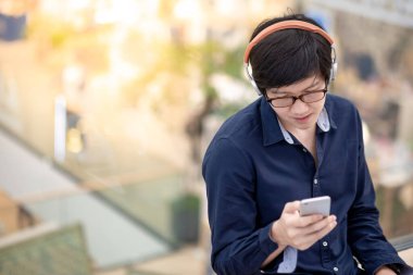 Asyalı bir adam alışveriş merkezinde dinlenirken müzik dinlerken akıllı telefon ve kulaklık kullanıyor. Cihaz teknolojisi ile modern yaşam tarzı.