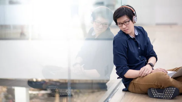 Asian man in casual clothing listening to music and looking around while sitting on the floor with laptop computer in public building.  Urban lifestyle concept