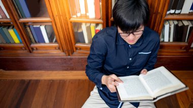 Akıllı Asyalı üniversite öğrencisi klasik kitaplığın yanında oturmuş kitap okuyor. Eğitim ve araştırma için üniversite kütüphanesinde ders kitabı kaynakları. Eğitim fırsatı için burs.