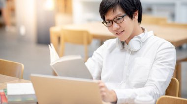Kolej kütüphanesinde online ders için dizüstü bilgisayar kullanırken gözlük okuyan Asyalı genç bir üniversite öğrencisi. E-öğrenme ve internette araştırma yapma. Eğitim kavramı