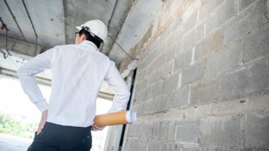 Asyalı inşaat mühendisi ya da koruma kaskı takan inşaat işçisi elinde yuvarlanmış planlar tutuyor. İnşaat alanında çalışan erkek mimar. İnşaat ve mimari tasarım kavramları