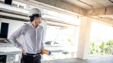 Asyalı inşaat mühendisi ya da koruma kaskı takan inşaat işçisi elinde yuvarlanmış planlar tutuyor. İnşaat alanında çalışan erkek mimar. İnşaat ve mimari tasarım kavramları