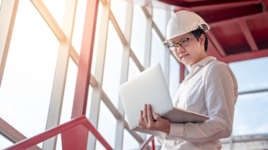 Asyalı inşaat mühendisi ya da miğfer takan inşaat işçisi dizüstü bilgisayar tutuyor. Erkek mimar inşaat alanında kırmızı merdivende çalışıyor. İnşaat ve mimari kavramı