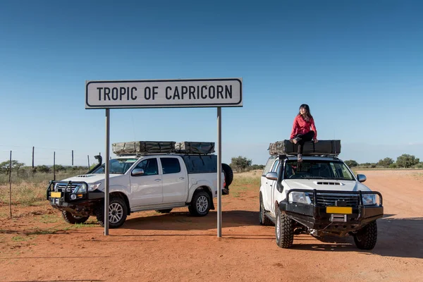 Asian woman traveler posing on camper car at tropic of capricorn sign in Namibia. Road trip travel in Africa concept.