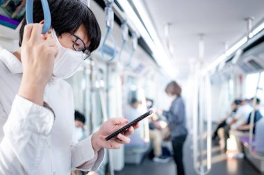 Asyalı bir adam hava treninde ya da şehir treninde akıllı telefon kullanarak cerrahi maske takıyor. Coronavirus (COVID-19) toplu taşımacılıkta salgın önleme. Pandemik koruma için sosyal uzaklık