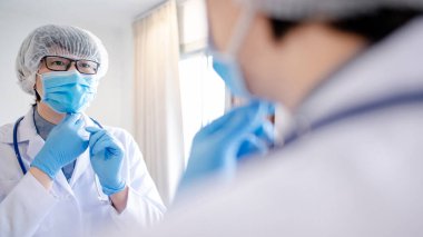 Asyalı bir erkek doktor ya da tek kullanımlık şapka takan bir doktor, cerrahi maske ve aynada kendine bakan lateks eldivenler. Tıbbi pratisyen, hastane kliniğinde muayene için hazırlan..