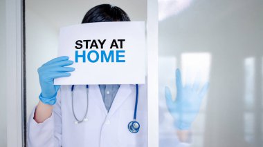 Kişisel koruyucu ekipman (PPE) giysisi, maskesi ve eldiveni olan doktor ya da tıp çalışanı üzerinde mesaj yazan kağıt olan evde kal. Coronavirus (COVID-19) salgını sırasında karantina kampanyası.