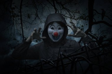 Scary evil clown wear jacket with old fence over dead tree clipart