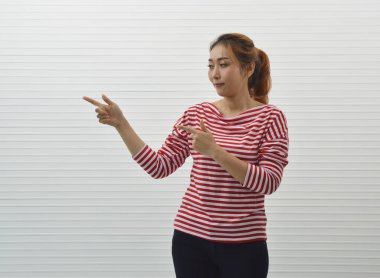 Mutlu genç asyalı kadın kırmızı ve beyaz soyulmuş gömlek ve kot giyen beyaz duvar arka plan üzerinde duran bir şey sunmak için parmak işaret, Gülümseyen yüz ifadesi, İş sunum konsepti