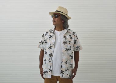 Hasır şapka, güneş gözlüğü, yaz gömleği ve kahverengi şort giyen özgüvenli gezgin Asyalı adam beyaz duvar arkasında duruyor, iş yazı tatili konsepti