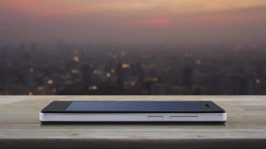 Günbatımında şehir manzarası bulanıklığı üzerine ahşap masadaki modern akıllı cep telefonu ekranında düğmeli düz simge oynat, iş müziği online konsepti