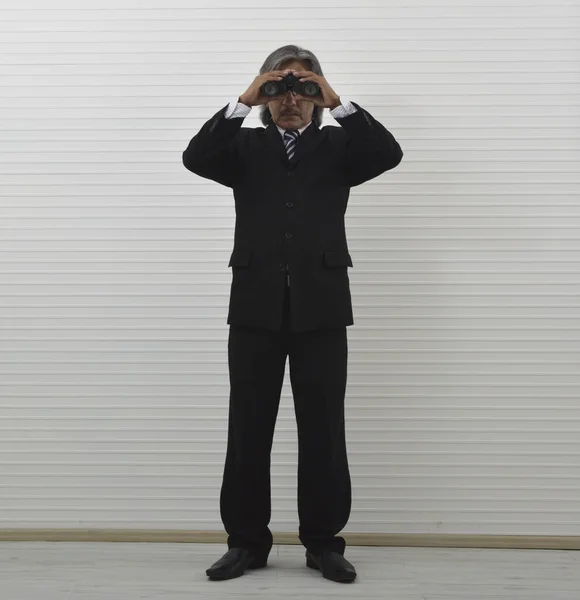 Elderly asian businessman in black suit and eyeglasses holding binoculars standing over white wall background, Business vision or marketing research concept