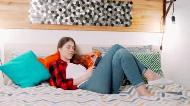 Genç ve çekici beyaz bir kadın, modern yatak odasında yatağında uzanmış elinde tuttuğu akıllı telefona bağlı Airpod 'lar gibi modern kulaklıkları dinlerken gülümsüyor.