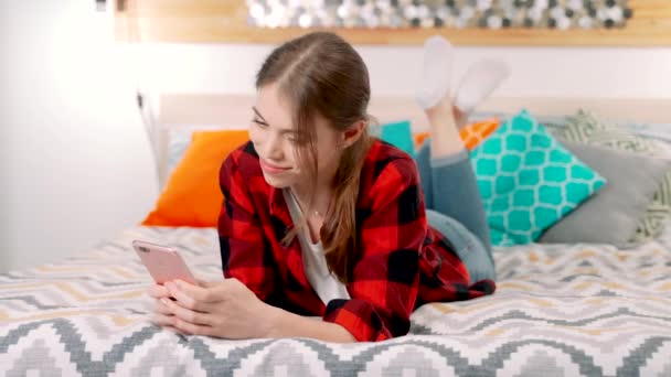 一位年轻貌美的白人妇女微笑着躺在床上 手里拿着一部手机 与她的朋友或亲戚发短信 — 图库视频影像