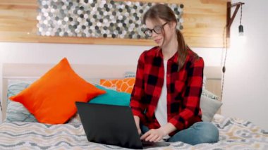 Genç bir öğretmen, öğrenci ya da yazar olabilecek genç bir kadın laptopta çalışıyor. Çift kişilik yatakta oturuyor ve gülümsüyor.